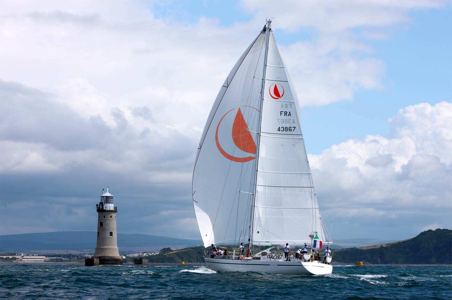 A Yacht sails fast towards a lighthouse, it's a cloudy day but the sun still shines making the white of the sails pop!