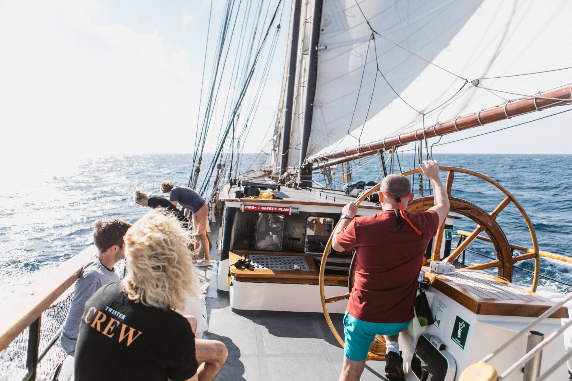 Looking down a ship in sail! A man stands holding a helm as big as him as the rest of the crew sit to the side enjoying the wind and calm seas