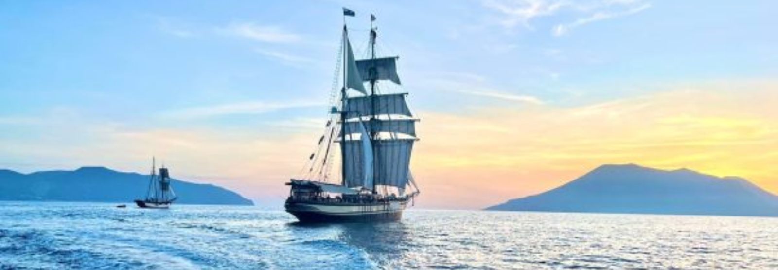 A tall ship with their sails out wide sailing into a hazy sunset or sun rise. Its a photo of cool pastels and calm seas