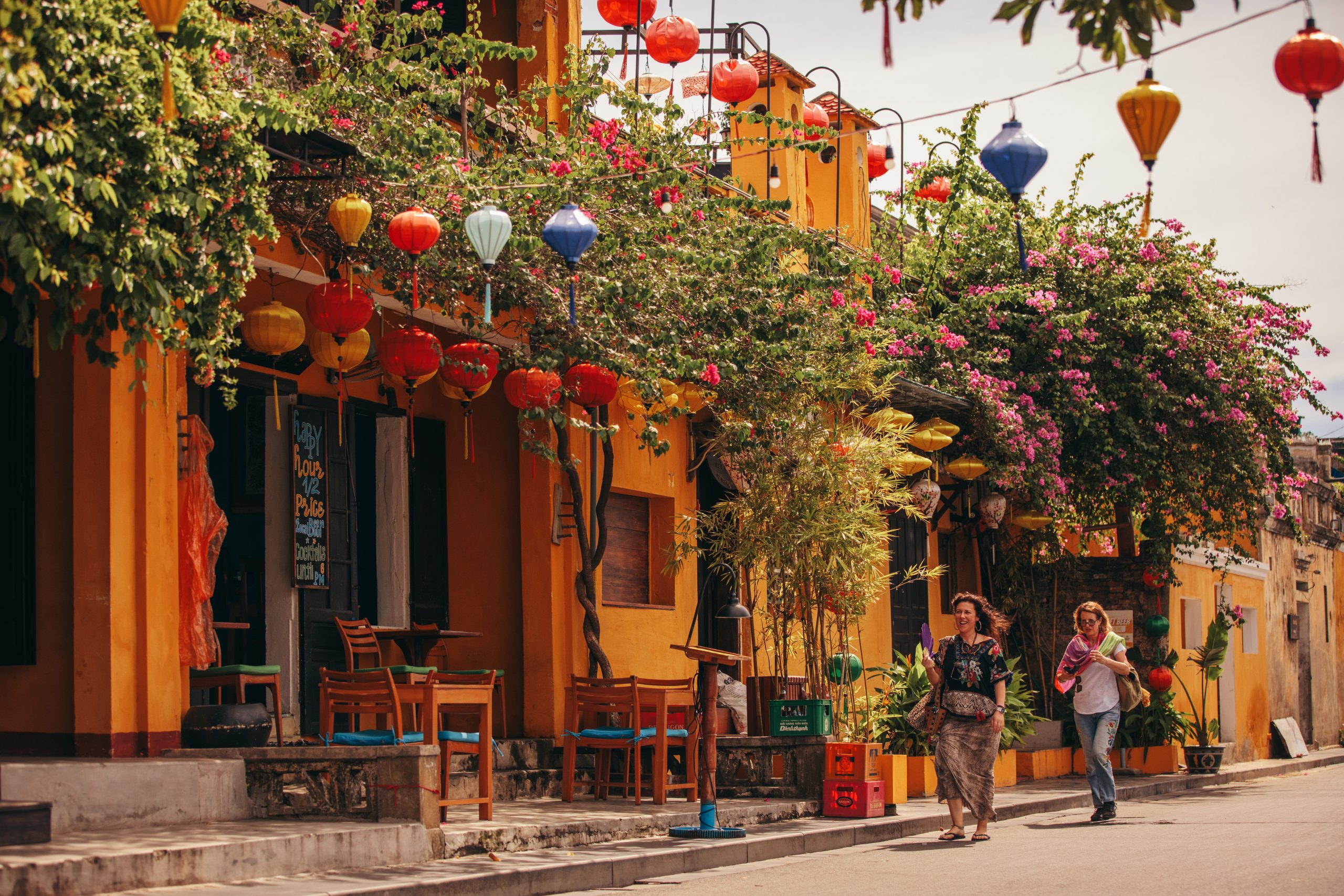 Two people walk through a vibrant street covered in plants and lanterns